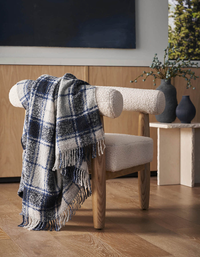 A white sherpa chair with wooden legs features the Cozy Earth Alpaca Boucle Throw draped on its back. A vase with greenery sits on a light-colored side table behind, and the setting rests on a wooden floor.|Color:Indigo Plaid
