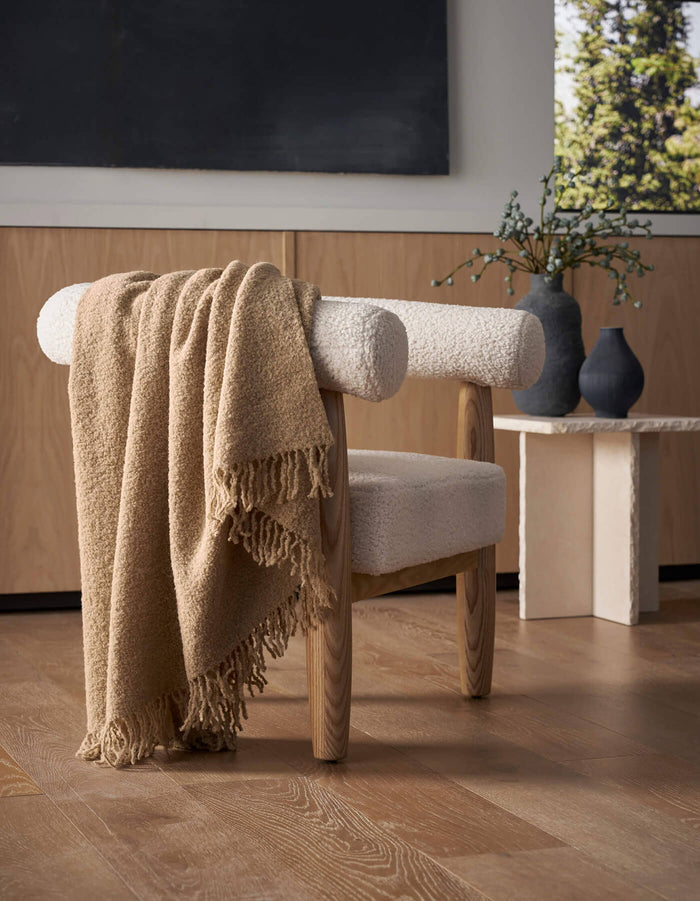 A modern wooden chair with white upholstery sits on a light wood floor, draped with the Cozy Earth Alpaca Boucle Throw. In the background, a small table displays two matte vases with greenery beside a window revealing trees outside.|Color:Sandalwood