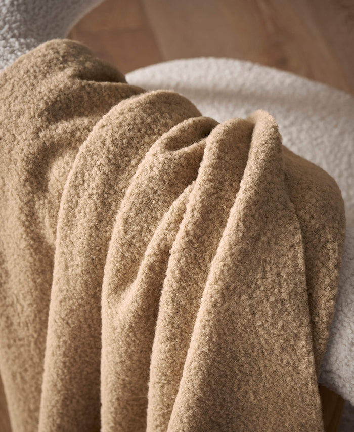 A close-up of the Cozy Earth Alpaca Boucle Throw in soft beige, elegantly draped over a white fuzzy chair, with a wooden floor in the background.|Color:Sandalwood