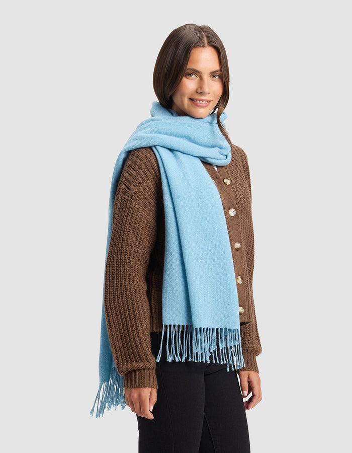 A woman with shoulder-length brown hair smiles while standing against a plain light gray background, wearing Cozy Earth's Amelia Wrap in brown over black pants and a light blue fringed scarf draped around her neck.|Color:Light Blue