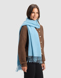 A woman with shoulder-length brown hair smiles while standing against a plain light gray background, wearing Cozy Earth's Amelia Wrap in brown over black pants and a light blue fringed scarf draped around her neck.|Color:Light Blue