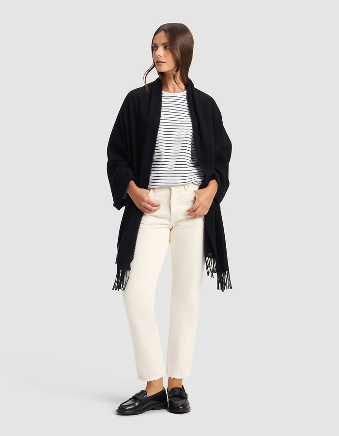 A woman stands against a plain background, wearing the Cozy Earth Amelia Wrap—a black shawl with fringe—over a white and black striped shirt, off-white pants, and black loafers. She looks to the side with her hands in her pockets.|Color:Black