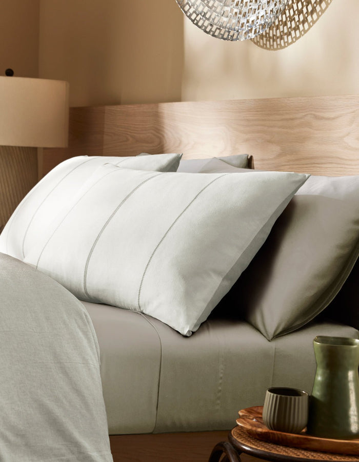 A modern bedroom featuring a neatly made bed with Cozy Earth’s Baja Double Sided Sham, light gray pillows and sheets, a wooden headboard, a bedside table holding a green cup and saucer, and a lamp under soft lighting.|Color:Laurel Stripe/Striped Bone