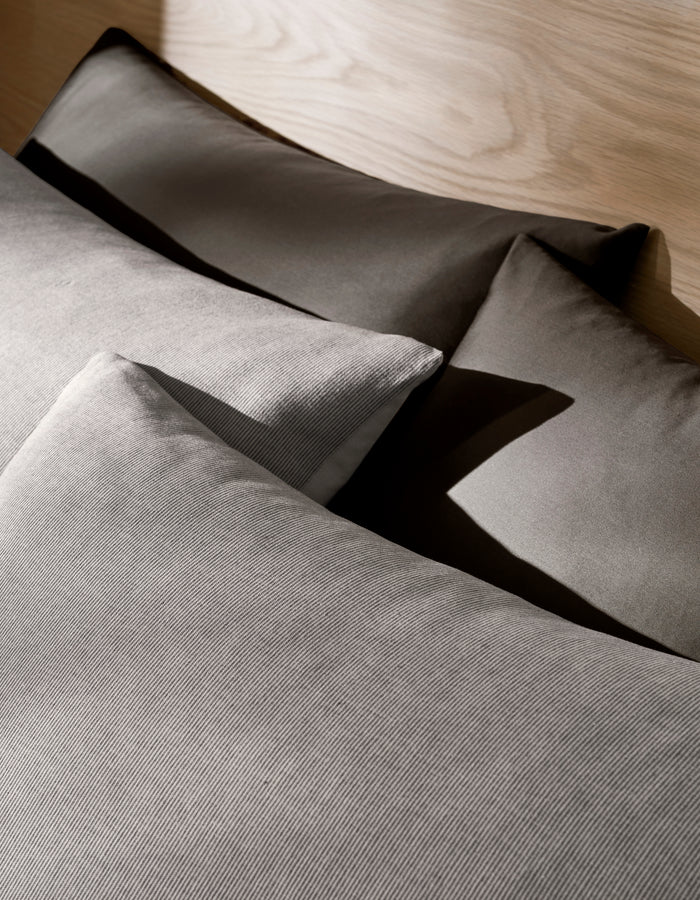 Four neatly arranged Baja Double Sided Shams by Cozy Earth rest on a bed with a light wooden headboard, creating soft shadows and a cozy, minimalistic atmosphere.|Color:Coal Stripe/Striped Alabaster