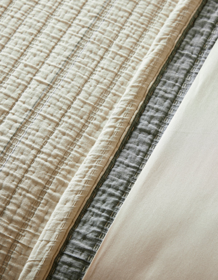 A close-up of layered bedding featuring the Cozy Earth Baja Matelasse Coverlet in beige, a gray quilted blanket, and a smooth cream-colored sheet.|Color:Alabaster/Oceanside