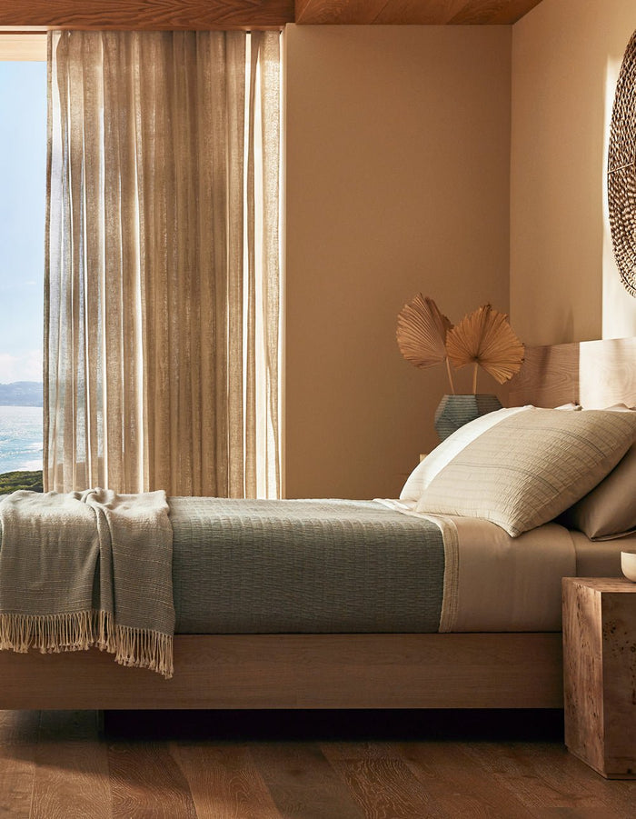 A sunlit, minimalist bedroom featuring the Cozy Earth Baja Matelasse Coverlet on a wooden bed, with neutral decor, a woven wall hanging, nightstand, dried leaves, and sheer curtains framing a sea view.|Color:Alabaster/Oceanside