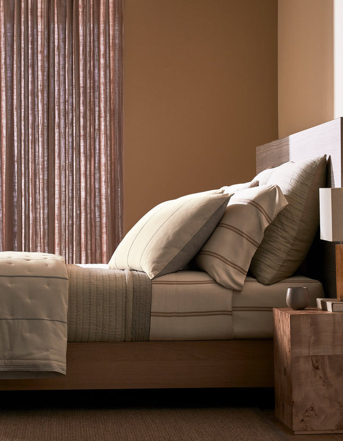 A neatly made bed featuring Cozy Earth's Baja Matelasse Bundle sits beside a wooden nightstand with a lamp and cup, set in a softly lit room with sheer curtains and beige walls.|Color:Ebony/Taupe