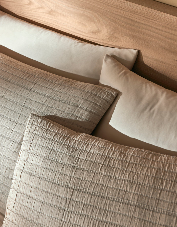 Two Cozy Earth Baja Matelasse Shams in beige and two plain cream pillows are arranged on a light wooden headboard, creating a cozy and inviting bed setting.|Color:Alabaster/Oceanside