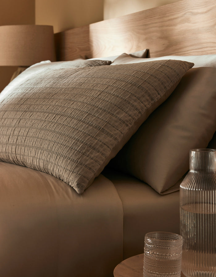 Close-up of a bed featuring Cozy Earth's Baja Matelasse Sham, with neutral bedding, pillows, a wooden headboard, and a glass carafe with tumbler on the bedside table. Warm lighting adds to the cozy atmosphere.|Color:Alabaster/Oceanside