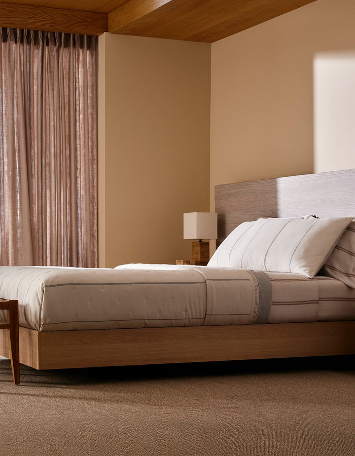 A modern bedroom features Cozy Earth’s Baja Quilt Bundle on a wooden platform bed, paired with a matching nightstand and lamp. Sheer curtains let in soft natural light, fostering a calm, minimalistic vibe.|Color:Coal Stripe/Striped Alabaster