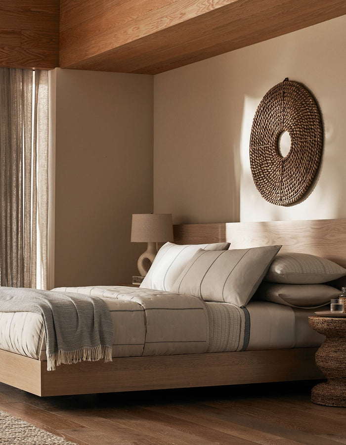 A modern, minimalist bedroom features a wooden bed frame with beige bedding, the Cozy Earth Baja Quilt, neutral pillows, a textured blanket, plus a woven circular wall decor, lamp, and round side table for a cozy sunlit look.|Color:Coal Stripe/Striped Alabaster