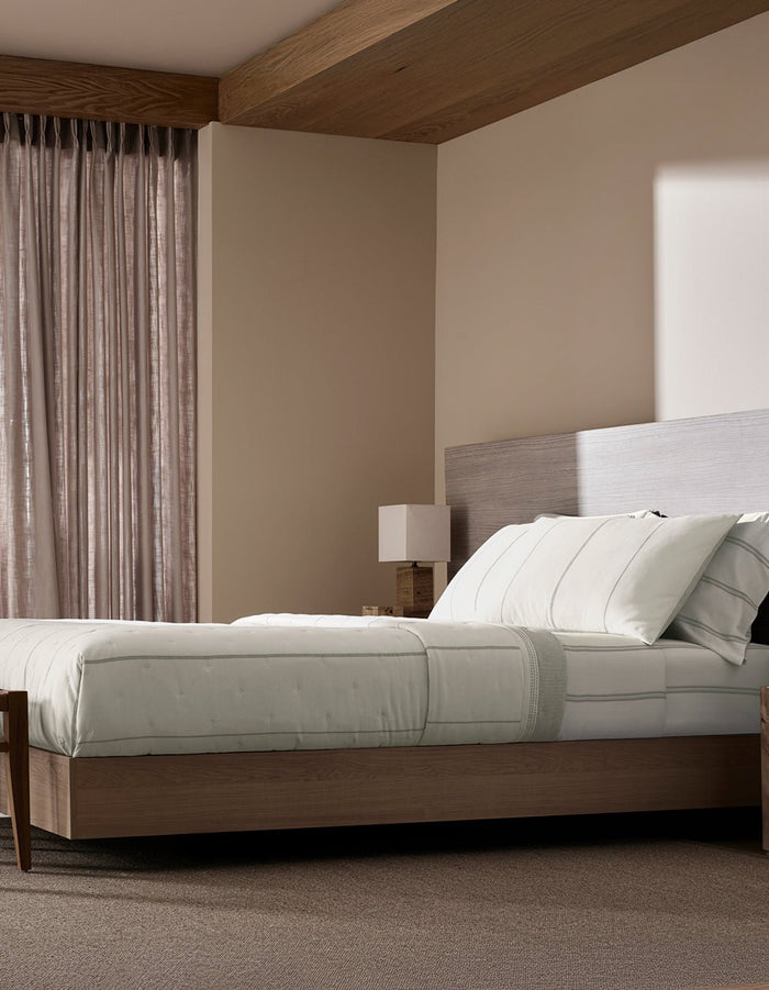 A modern bedroom with neutral tones features a wooden platform bed topped with the Cozy Earth Baja Quilt, light bedding, a minimalist nightstand with a square lamp, beige walls, and sheer curtains filtering sunlight that casts shadows on the wall.|Color:Laurel Stripe/Striped Bone