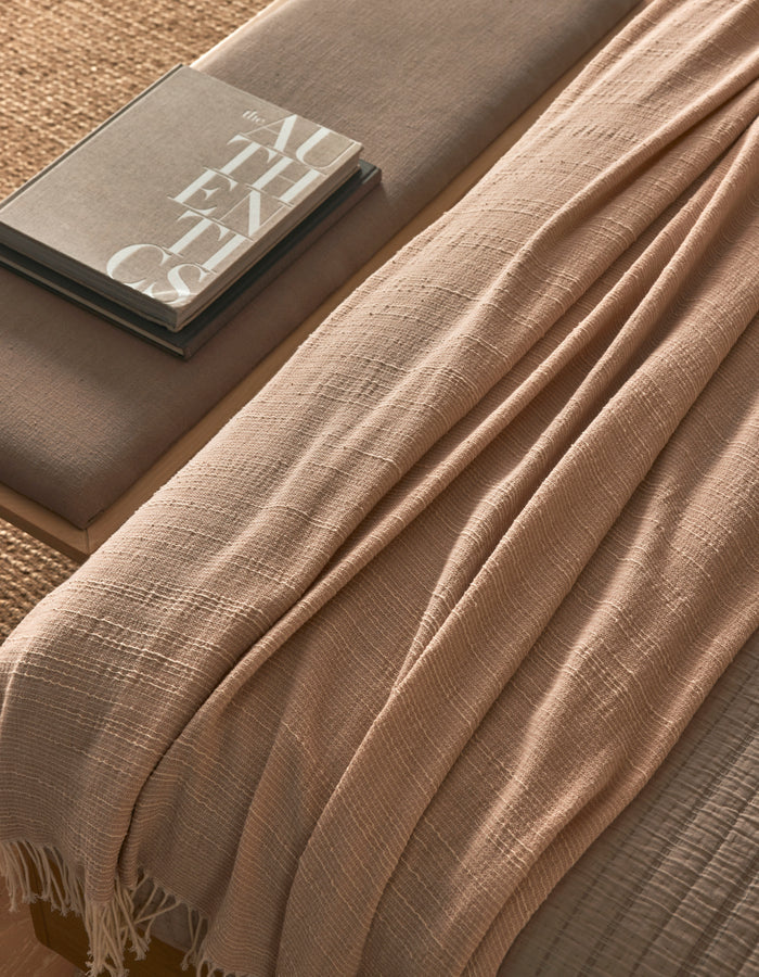 The Baja Tassel Throw by Cozy Earth, in beige with a textured finish, is draped over a neutral-toned bench beside two stacked books (one titled "AUTHENTICS"), creating a cozy, minimalistic look.|Color:Himalayan Salt