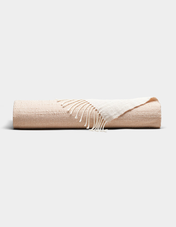 The Cozy Earth Baja Tassel Throw is a neatly rolled beige blanket with white fringed edges, showcased on a plain light gray background.|Color:Himalayan Salt