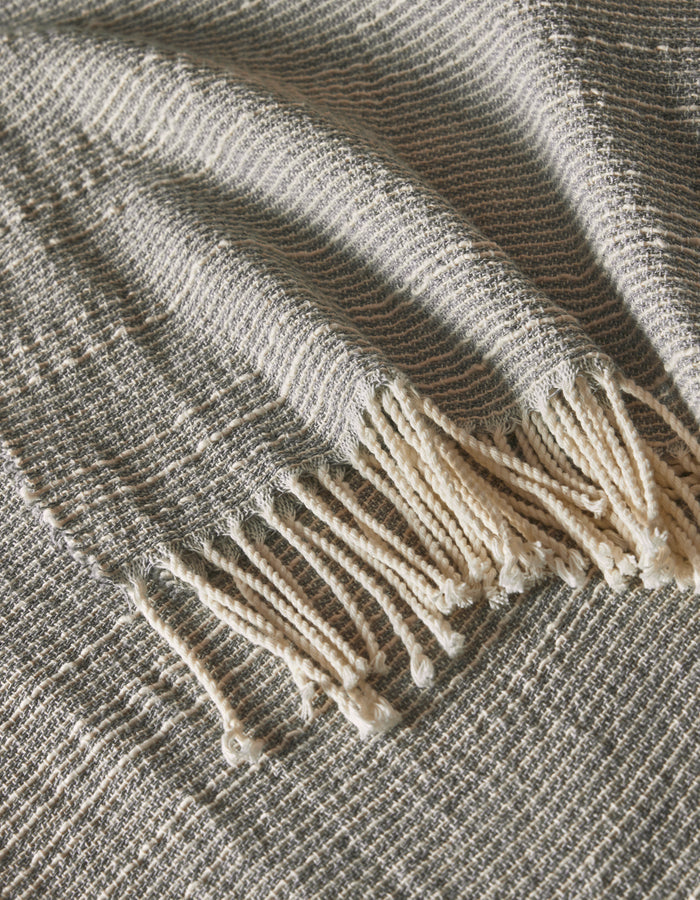 The Baja Tassel Throw by Cozy Earth is shown in a close-up, highlighting its gray and white textured weave and fringed tassels draped elegantly to display its detailed pattern.|Color:Grey