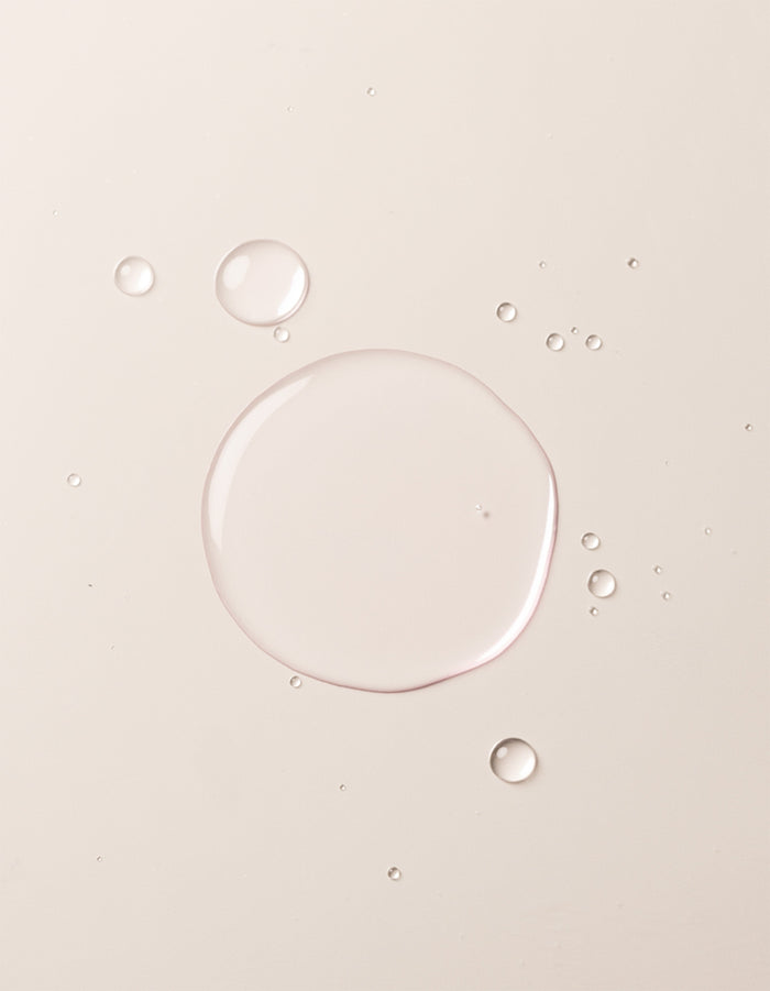 A close-up image showcases translucent liquid droplets of Cozy Earth's Balancing Face Mist on a smooth, off-white surface. The droplets vary in size, with one large central droplet surrounded by several smaller ones, creating a clean and minimalistic appearance. |Size:Full Size