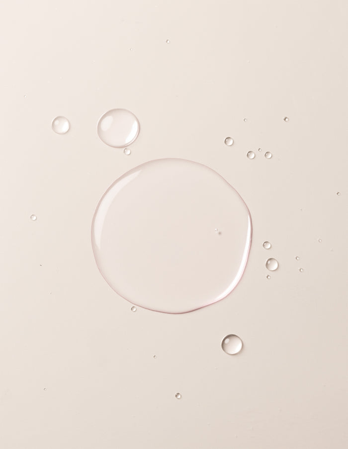 Clear liquid droplets of Balancing Face Mist by Cozy Earth are scattered on a smooth, light beige surface. The largest droplet is centered, surrounded by smaller drops, giving the image a minimal, clean, and soft appearance. |Size:Mini