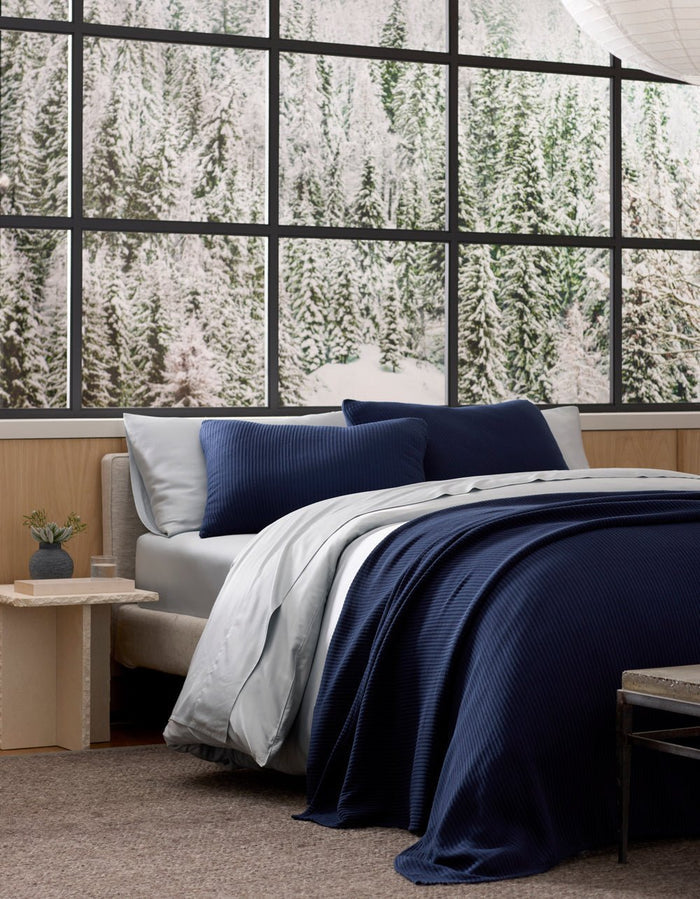 A neatly made bed featuring Cozy Earth's Bamboo Coverlet Shams in navy blue and crisp white sheets is beside a small nightstand. Large windows behind it showcase a snowy pine forest.|Color:Navy