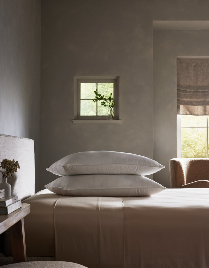 A serene bedroom with neutral tones features a neatly made bed topped with Cozy Earth Bamboo Down Alternative Pillows, a small window with a potted plant, and soft natural light streaming in from a larger window with a beige shade.Standard King