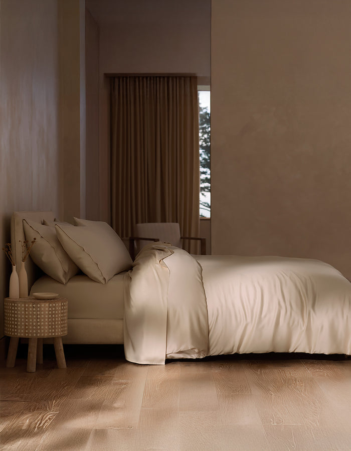 A minimal, modern bedroom features a neatly made bed with Cozy Earth's Bamboo Duvet Cover in cream, a round nightstand with a vase of dried plants, wood floors, and a window with brown curtains partially revealing greenery outside.|Color: Driftwood