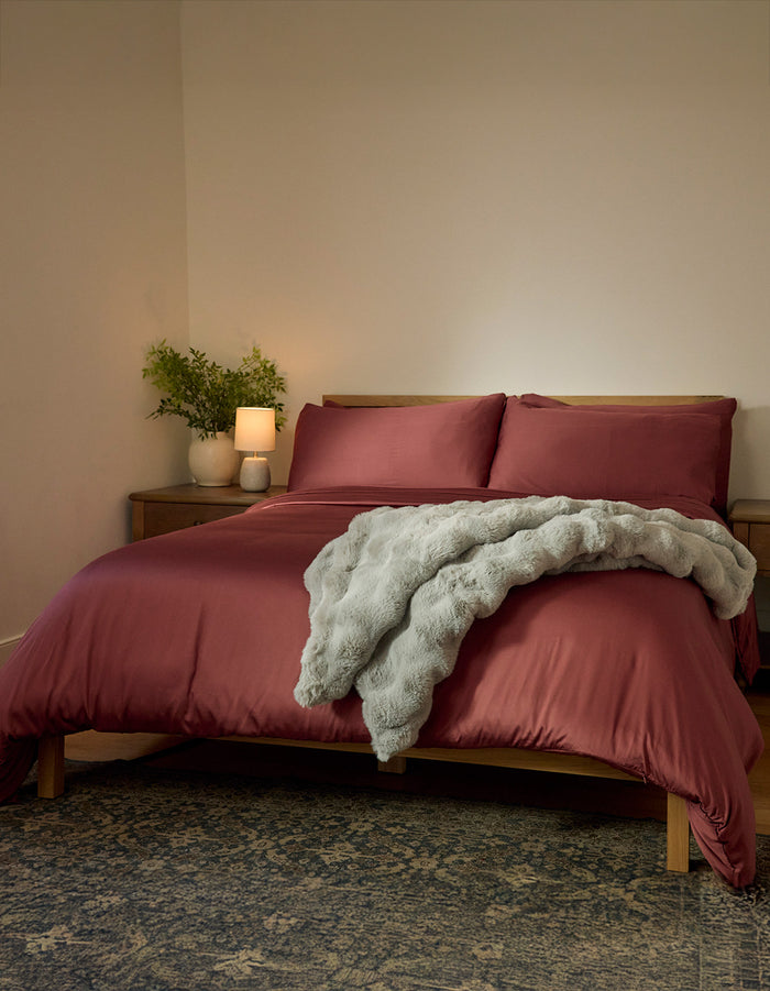 A neatly made bed features Cozy Earth's Bamboo Duvet Cover in dark red and a soft gray throw, set against a plain wall. A wooden nightstand with a plant, lamp, and candle sits beside the bed, with a patterned rug on the floor.|Color:Mulberry