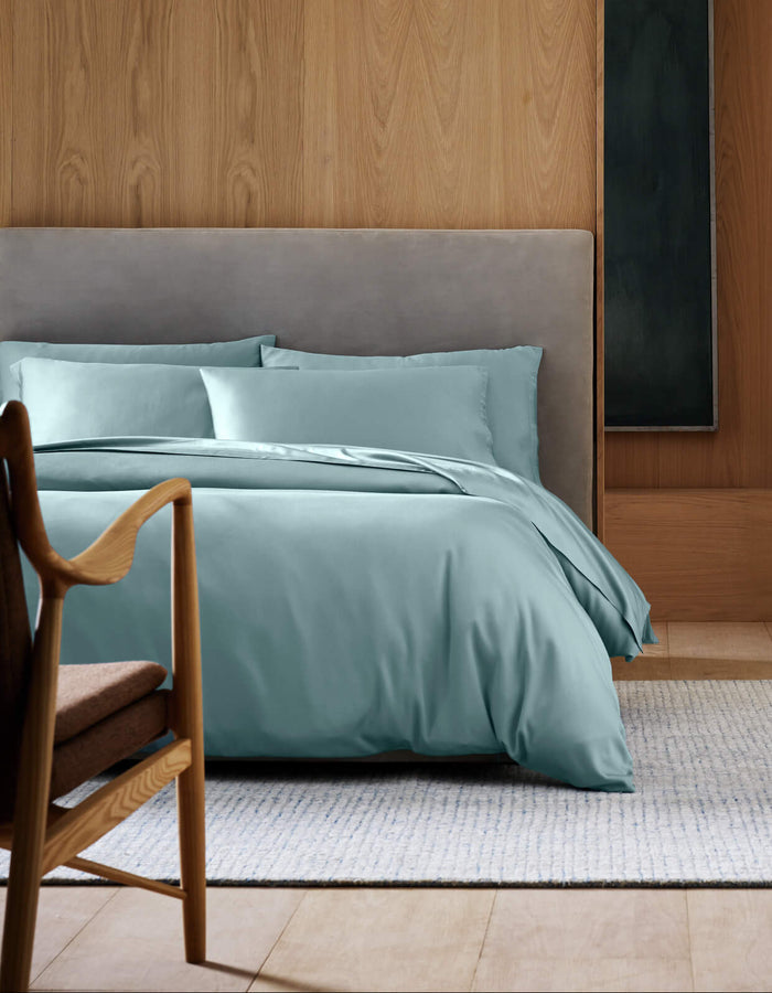 A modern bedroom featuring Cozy Earth's Bamboo Duvet Cover in light blue, a gray upholstered headboard, light wooden wall, wooden chair with brown cushion, and a light woven rug on a wooden floor.|Color:River
