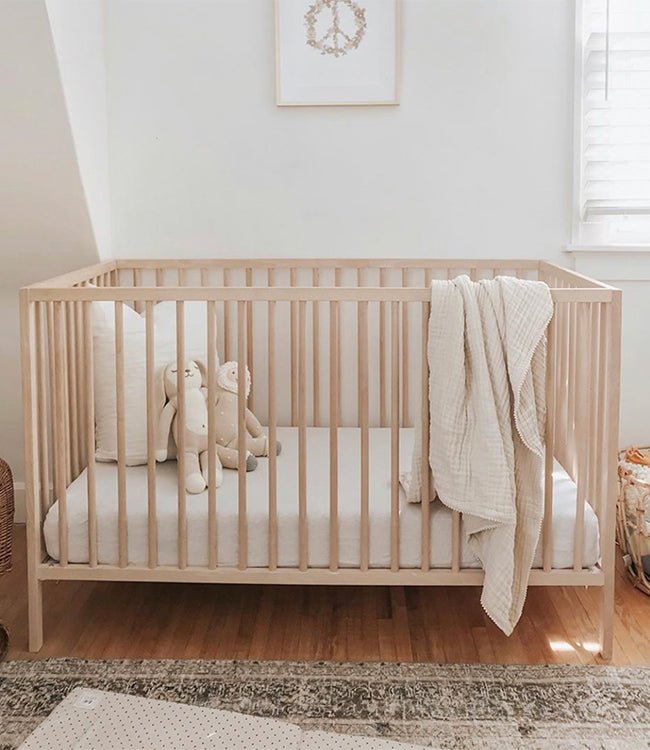 A minimalist nursery with a light wooden crib dressed in Cozy Earth’s Bamboo Fitted Sheet, white bedding, two stuffed animals, and a cream blanket draped over the side. The white-walled room features a window and framed print above the crib. |Color:Driftwood