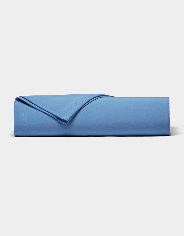 A neatly folded Bamboo Jersey Sheet Set by Cozy Earth in blue rests on a plain white surface, with one corner flipped to display the soft fabric texture.|Color:Sailor