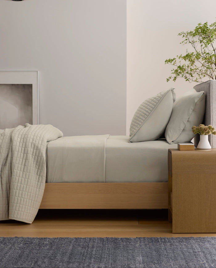 A minimalist room features a neatly made bed with Cozy Earth's Bamboo Jersey Flat Sheet in light gray, paired with pillows. Next to it is a wooden nightstand holding a small plant and vase, while a framed picture and pendant lamp complete the scene.|Color:Driftwood