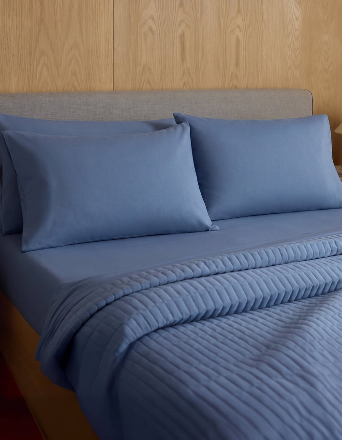 A neatly made bed features Cozy Earth Bamboo Jersey Pillowcases, matching pillows, and a quilted blue blanket, all set against a wood-paneled wall and a light gray upholstered headboard.|Color:Sailor