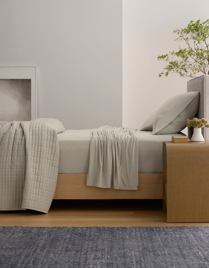 A minimal bedroom featuring Cozy Earth's Bamboo Jersey Sheet Set, a wooden bed, matching quilt, nightstand with plants, modern white pendant lamp, framed artwork on a light wall, and a blue rug on the floor.|Color:Driftwood