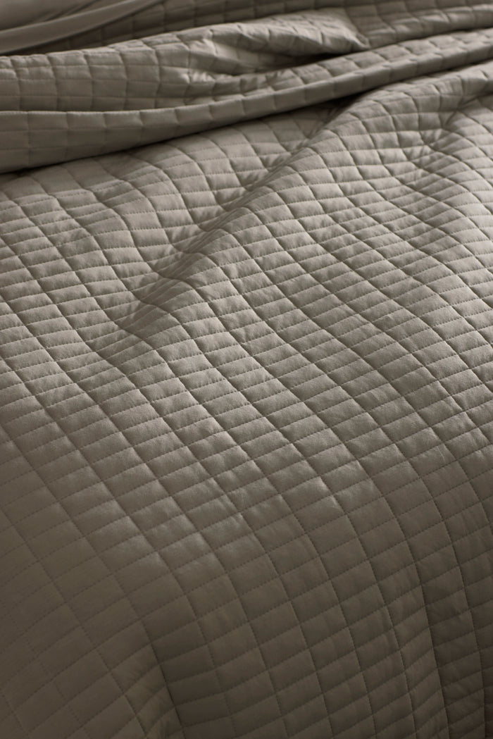 Close-up of a neatly made bed with the Cozy Earth Bamboo Jersey Quilt Bundle, featuring a soft, slightly wrinkled quilt in a light neutral tone with a grid of stitched squares.