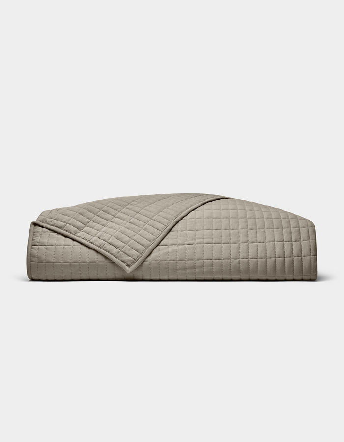 The Cozy Earth Bamboo Jersey Quilt is shown folded against a white background, with one corner turned down to display its beige color, grid pattern, and detailed stitching.|Color:Stone