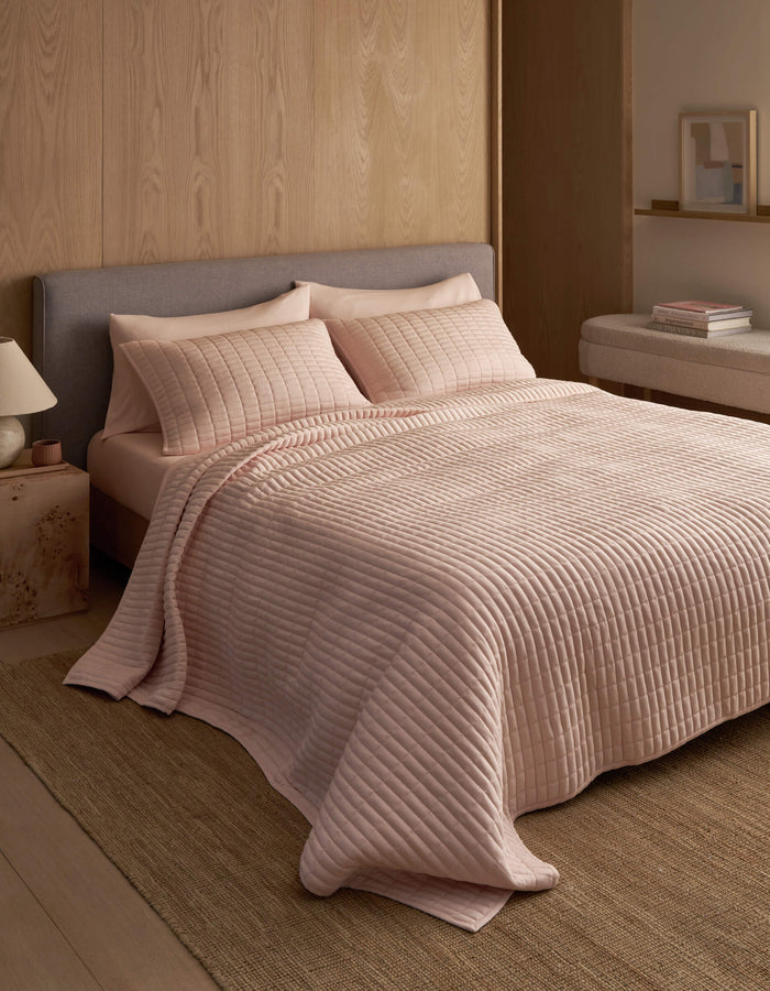A neatly made bed features the Cozy Earth Bamboo Jersey Quilt in light pink with matching pillows, set against a wooden wall in a minimal, modern bedroom with neutral tones and simple decor.|Color:Petal