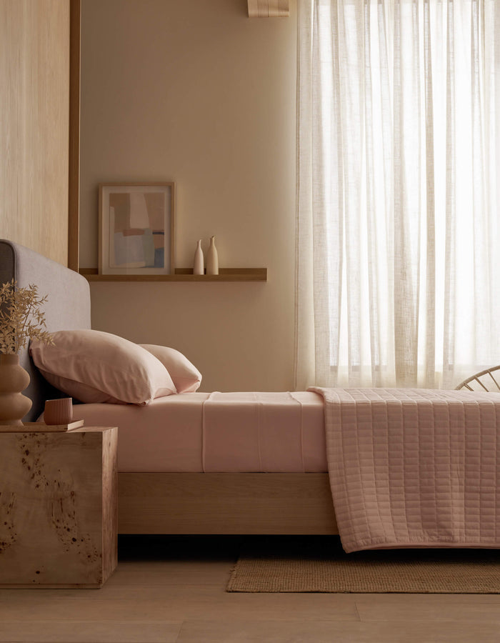 A minimalist bedroom features a neatly made bed with Cozy Earth Bamboo Jersey Flat Sheet in soft pink, a wooden nightstand holding a vase and candle, light wood walls, sheer curtains allowing natural light, and abstract art on a shelf.|Color:Petal