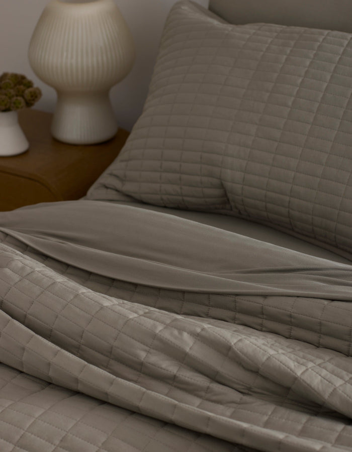 A neatly made bed features the Bamboo Jersey Quilt by Cozy Earth in taupe with matching pillows, next to a wooden nightstand topped with a white ribbed lamp and a small flower vase.|Color:Stone