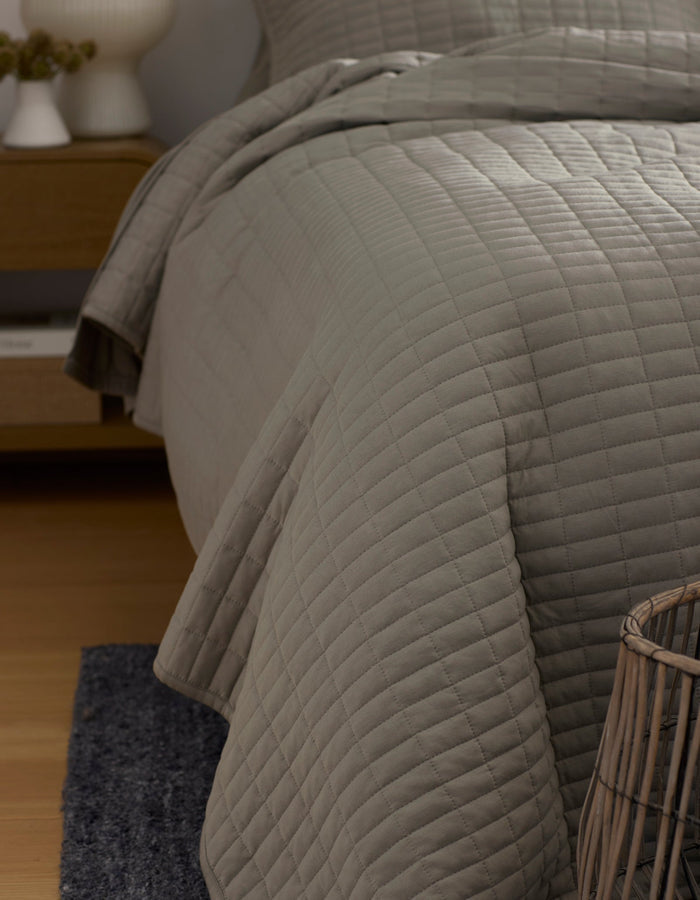 A close-up of a neatly made bed features the Cozy Earth Bamboo Jersey Quilt in light gray, draped over the side. Also visible are part of a wicker basket, a dark rug, and a wooden nightstand in this cozy bedroom corner.|Color:Stone