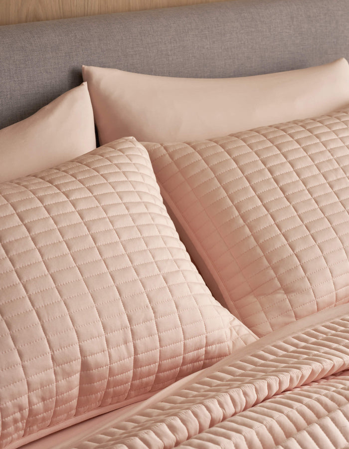 Close-up of a neatly made bed featuring Cozy Earth's Bamboo Jersey Quilted Shams and matching pillowcases in light pink, paired with a quilted bedspread. The upholstered headboard is covered in soft gray fabric.|Color:Petal