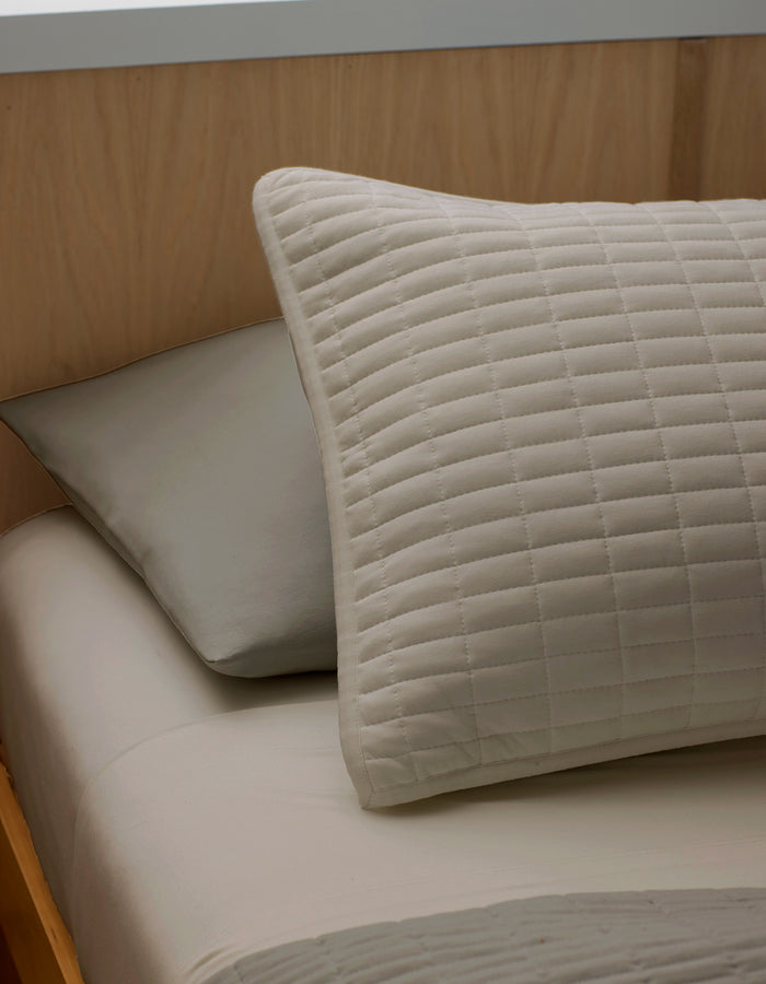 A close-up of a neatly made bed features two pillows resting against a light wooden headboard: a Bamboo Jersey Quilted Sham by Cozy Earth in beige in front, and a plain gray pillow behind.|Color:Driftwood