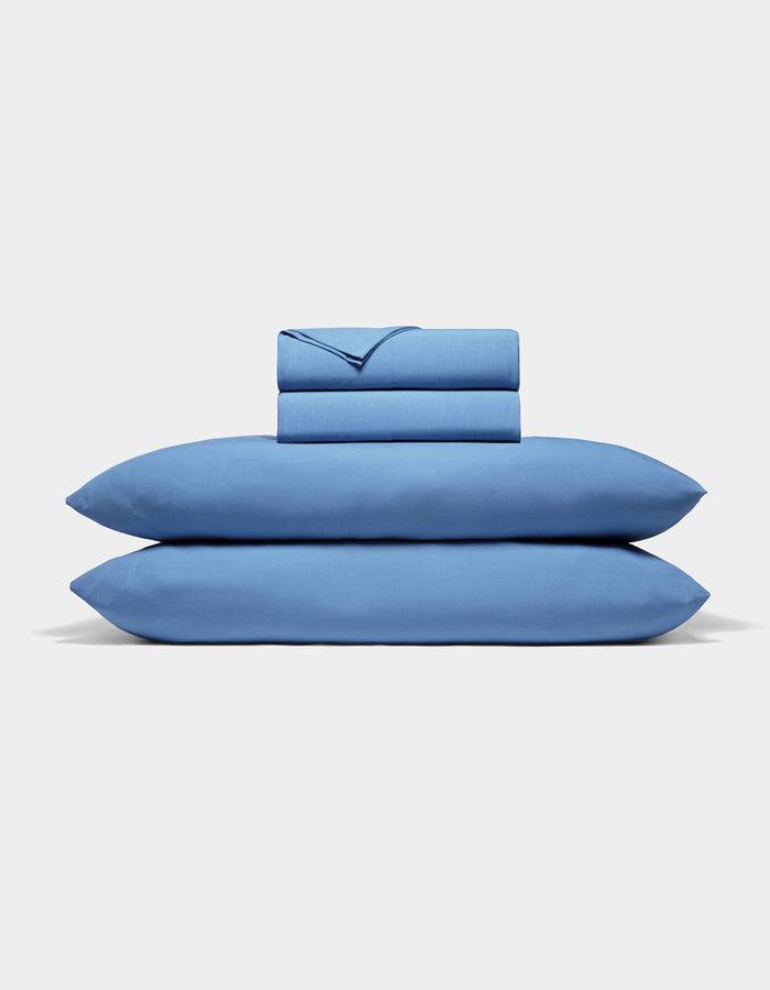 The Cozy Earth Bamboo Jersey Sheet Set, featuring two pillowcases, a fitted sheet, and a flat sheet in blue, is neatly folded and stacked against a plain white background.|Color:Sailor