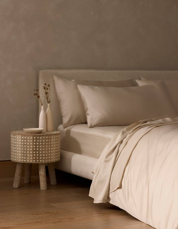 A minimalist bedroom features a beige upholstered bed dressed in Cozy Earth’s Bamboo Flat Sheet, accented with beige pillows. A round wooden nightstand holds two simple vases with dried flowers, complementing the warm, neutral color palette.|Color:Driftwood