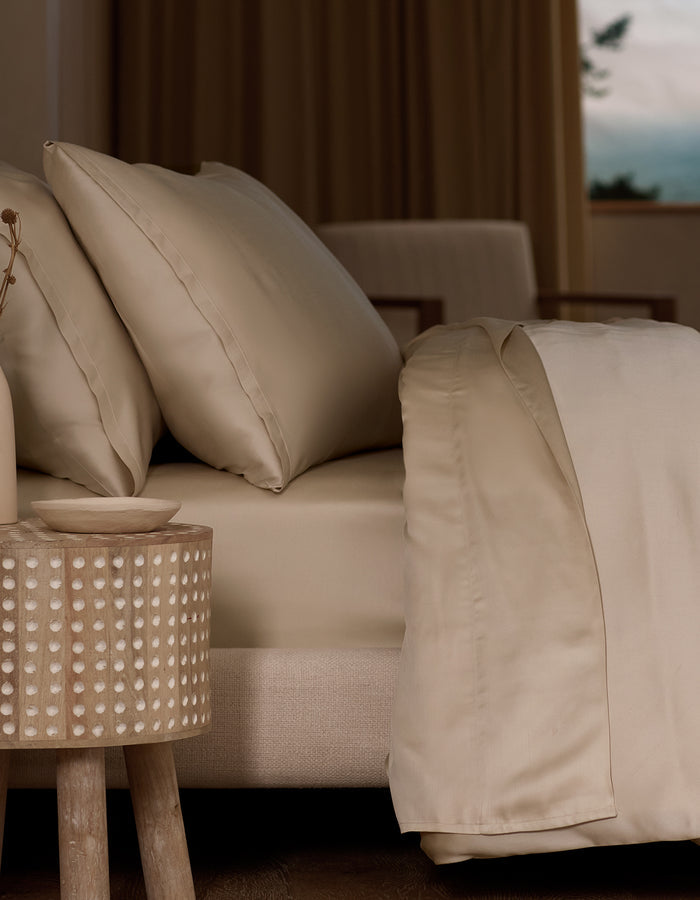 A neatly made bed features Cozy Earth Bamboo Pillowcases in beige, next to a round wooden bedside table with a dotted pattern in a softly lit, neutral room. A window in the background reveals an outdoor view.|Color:Driftwood