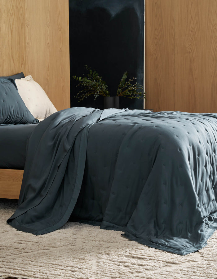 A neatly made bed features Cozy Earth's Bamboo Quilt in deep blue, paired with a textured blanket. Against a wooden wall, the room includes a light rug and a bedside table with a black vase of green foliage.|Color:Pacific Blue