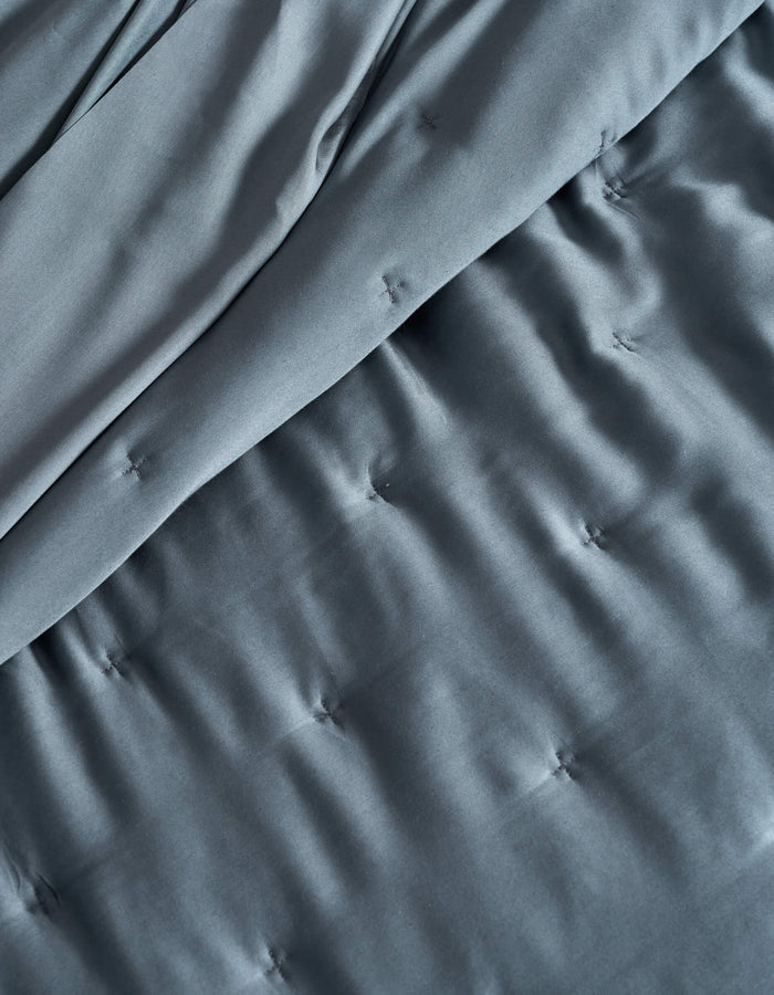 A close-up of the Cozy Earth Bamboo Quilt shows its dark gray, silky, and slightly shiny fabric with a quilted pattern, stitched tufts, and gentle folds.|Color:Pacific Blue