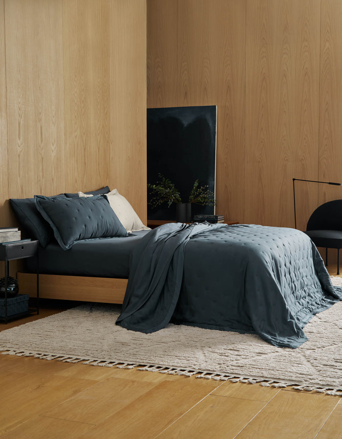 A modern bedroom with light wood walls and floor features a dark gray bed dressed in the Cozy Earth Bamboo Quilt, a small side table, light rug, black chair, and a dark abstract painting behind lush plants in a black vase.|Color:Pacific Blue