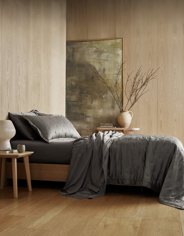 A modern bedroom with light wood walls and floors features a bed dressed in the Cozy Earth Bamboo Quilt in dark gray, a nightstand with a white lamp, another table with a vase of branches, and abstract artwork in the background.|Color:Charcoal