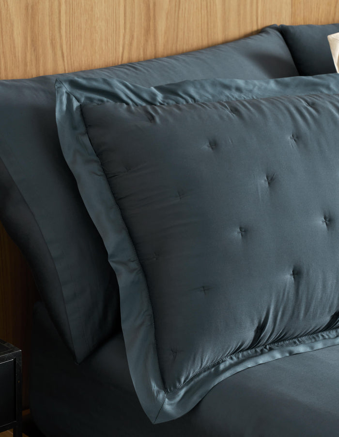 A close-up of a neatly made bed features Cozy Earth Bamboo Quilted Shams and matching sheets in dark blue, set against a light wood headboard.|Color:Pacific Blue