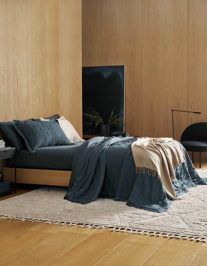 A modern bedroom features wood-paneled walls, a low bed with Cozy Earth's Bamboo Quilt Bundle in dark teal, a cream throw, and pillows. A neutral woven rug, black chair, side table, and minimalist decor create a cozy, stylish space.|Color:Pacific Blue