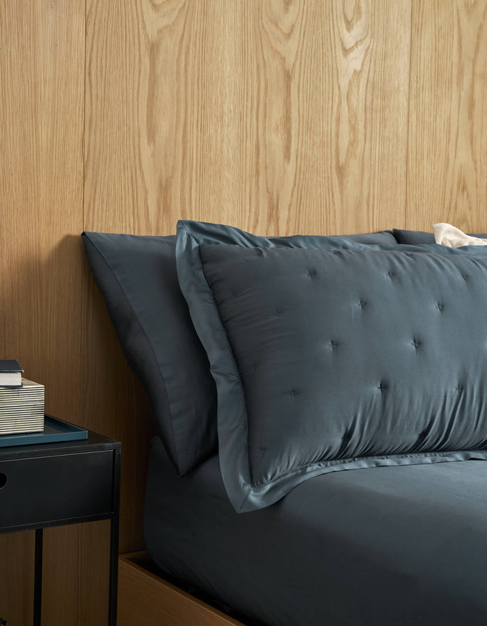 A neatly made bed with Cozy Earth Bamboo Quilted Shams in dark gray and matching sheets sits against a light wood-paneled wall. A black nightstand with stacked books is next to the bed.|Color:Pacific Blue
