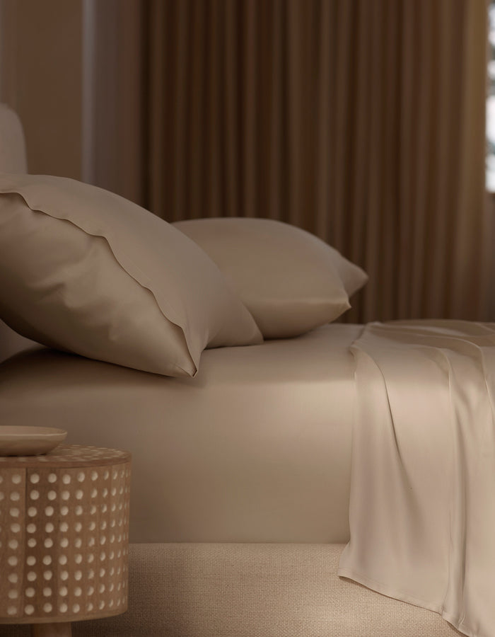 A neatly made bed features Cozy Earth’s Bamboo Sheet Set in beige with matching pillows, beside a round wooden nightstand with a dotted design. Soft, warm light through curtains enhances the cozy atmosphere. |Color:Driftwood